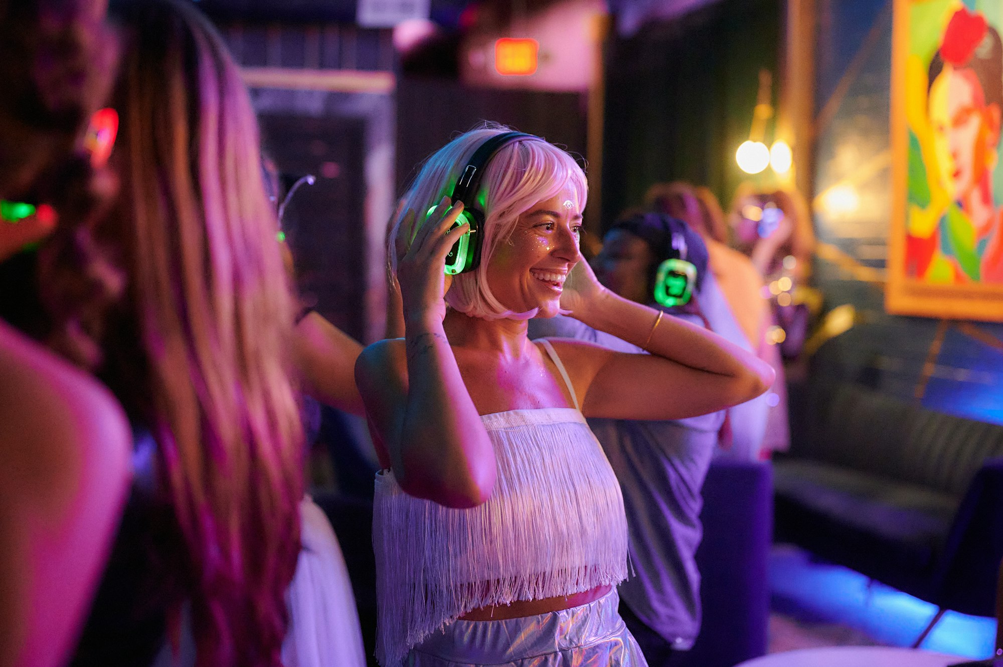 A woman smiling at a night silent disco, hands on glowing green wireless headphones